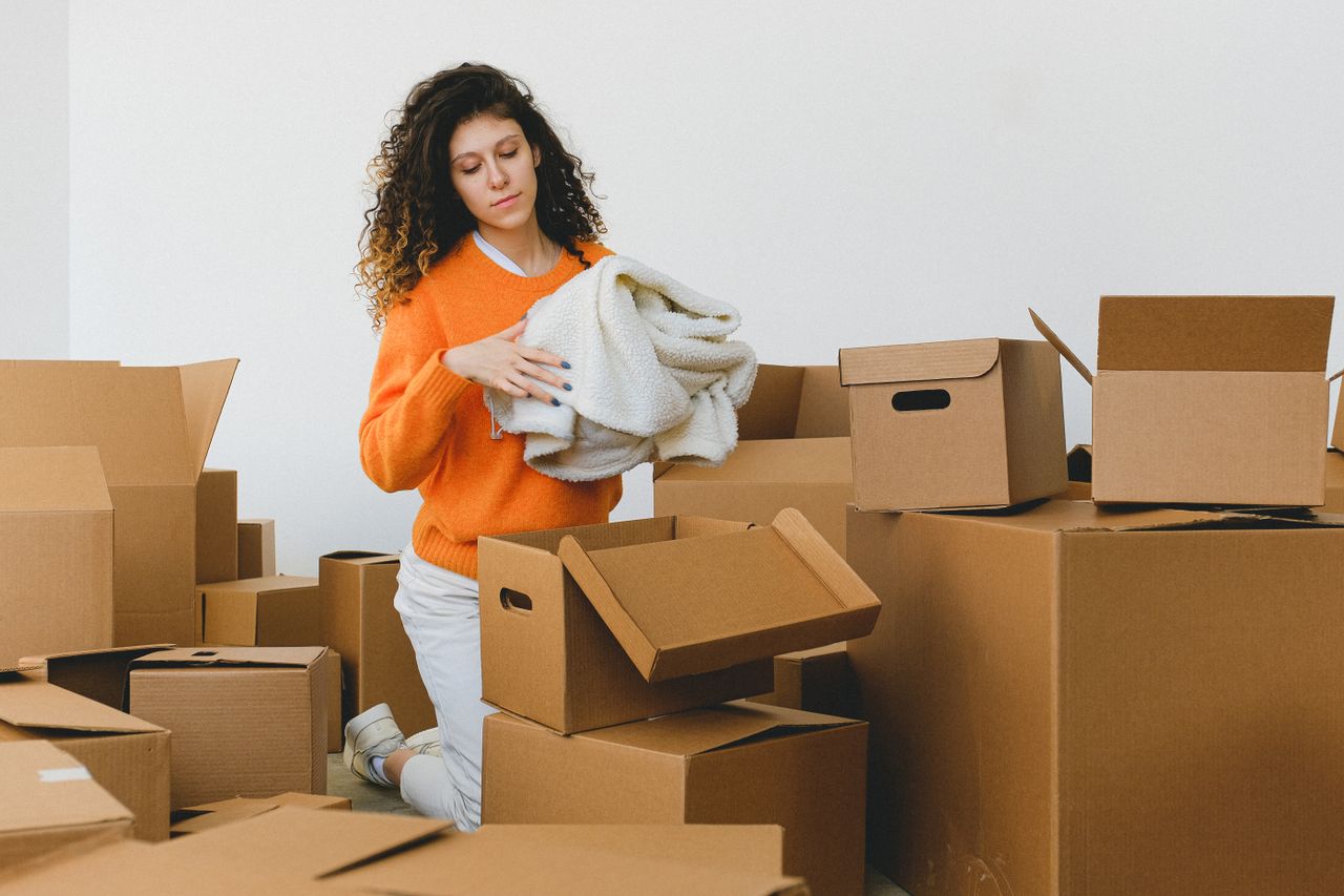 Jeune femme concentrée aux cheveux bouclés, agenouillée sur le sol et rangeant des vêtements dans des cartons avant de déménager dans son nouvel appartement